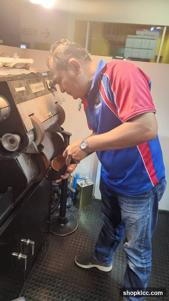 A man in a blue and red polo shirt and jeans is working on a shoe at a shoe repair machine. The machine has various polishing and buffing wheels. In the background, a sign reads "LOBBY" with an arrow pointing to the right.
