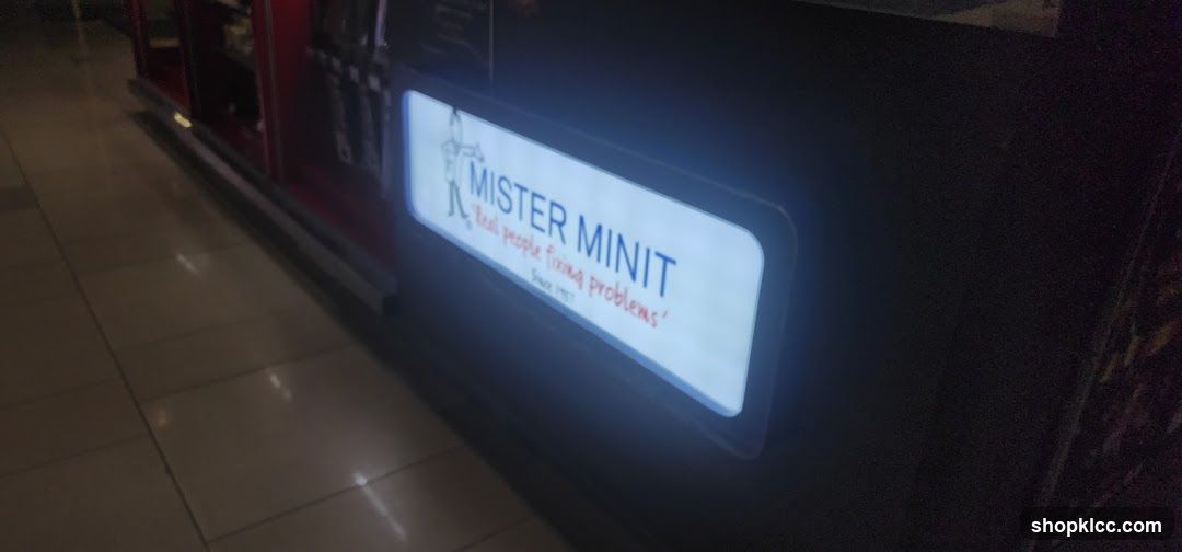 A lit sign with the Mister Minit logo and slogan. The logo features a stylized figure and the text reads "MISTER MINIT" in blue, with the slogan "Real people fixing problems" in red below it. The founding year "Since 1957" is also visible in small red text. The sign is mounted on a dark surface, possibly a counter or display case.