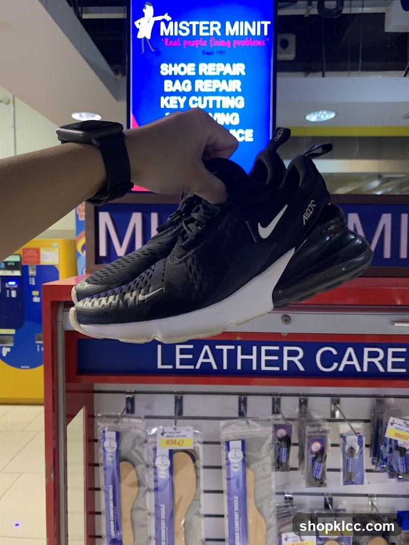 A hand holds a pair of black Nike sneakers in front of a Mister Minit store display. The display advertises shoe repair, bag repair, and key cutting services. Below the services list, there are shelves stocked with shoe care products, including insoles priced at RM47.