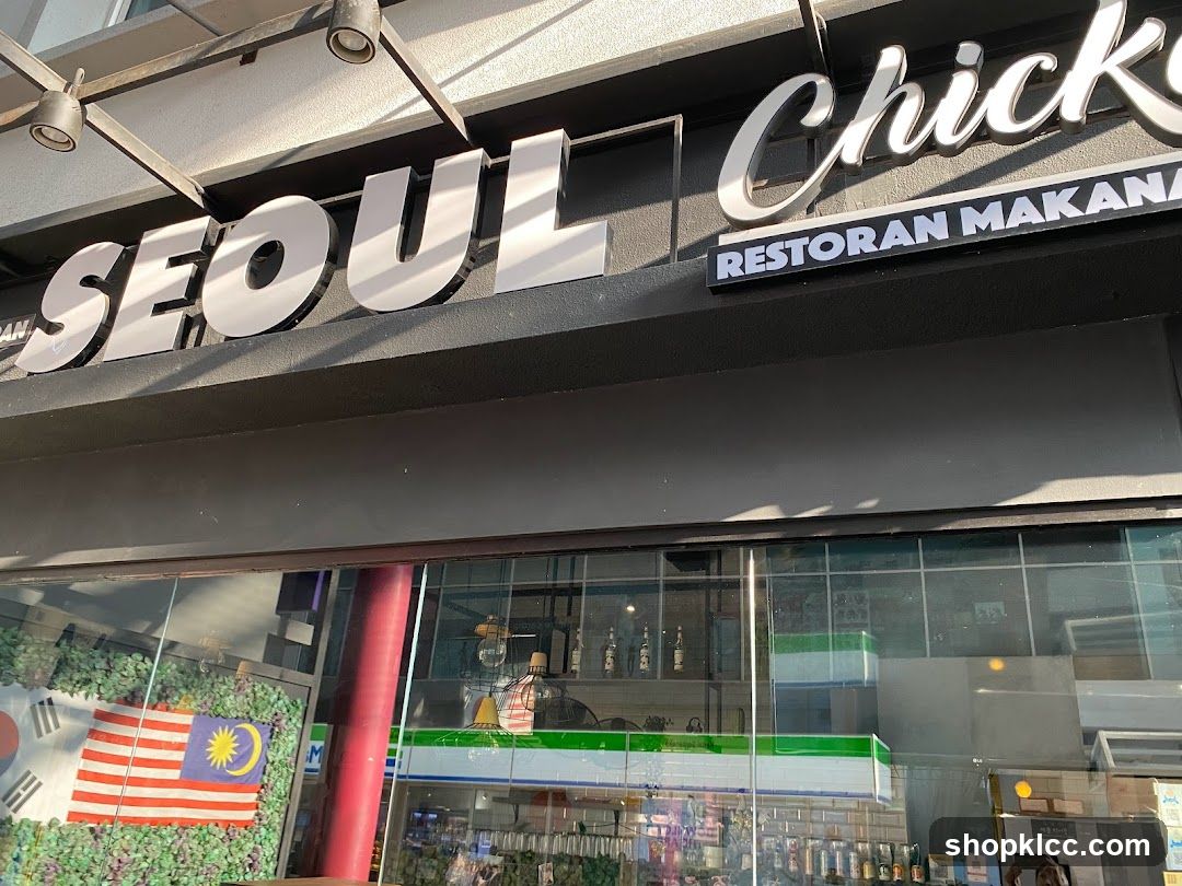 The exterior of a restaurant called Seoul Chicken. The large, white, three-dimensional letters spelling "SEOUL" are prominent above the entrance. To the right, smaller white letters on a black background read "Chicken" and below that, "RESTORAN MAKANAN". Through the glass storefront, flags of South Korea and Malaysia are visible, along with interior lighting and shelves with bottles.