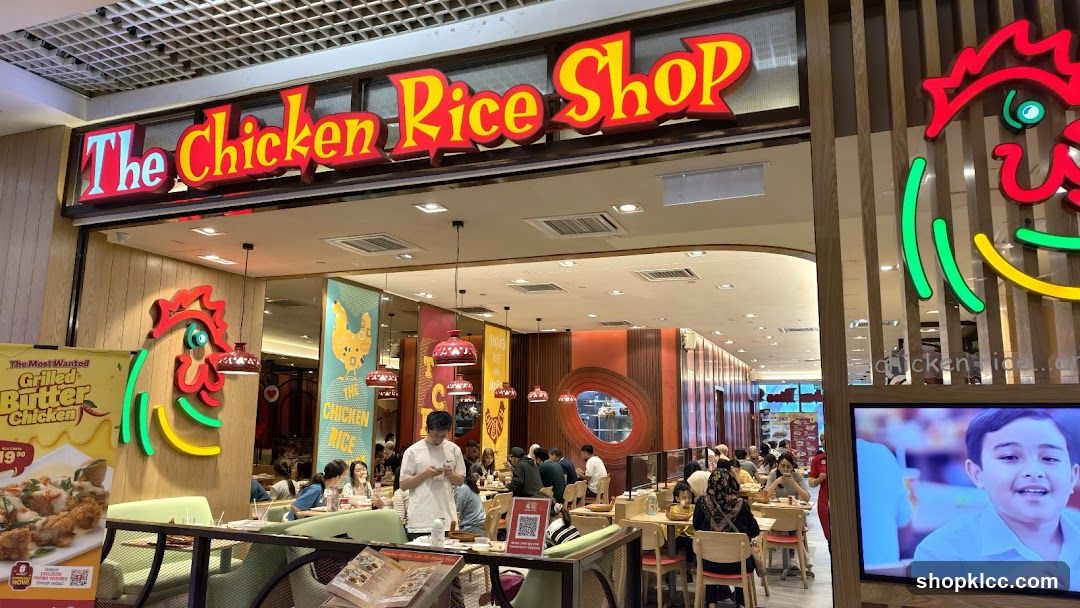The exterior of The Chicken Rice Shop restaurant. A large red and yellow sign with the shop's name is visible above the entrance. Inside, diners are seated at tables. A promotional poster for "The Most Wanted Grilled Butter Chicken" is displayed on the left.
