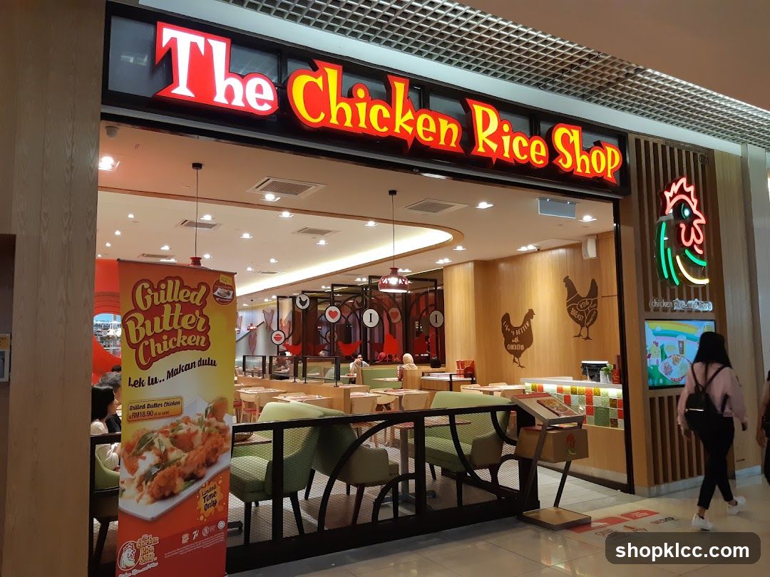 The exterior of a restaurant called "The Chicken Rice Shop". A large red and yellow sign with the restaurant's name is visible above the entrance. To the right of the entrance, a neon sign in the shape of a chicken is displayed. Inside the restaurant, tables and chairs are visible, along with wooden walls decorated with chicken silhouettes. A promotional banner for "Grilled Butter Chicken" is placed to the left of the entrance.