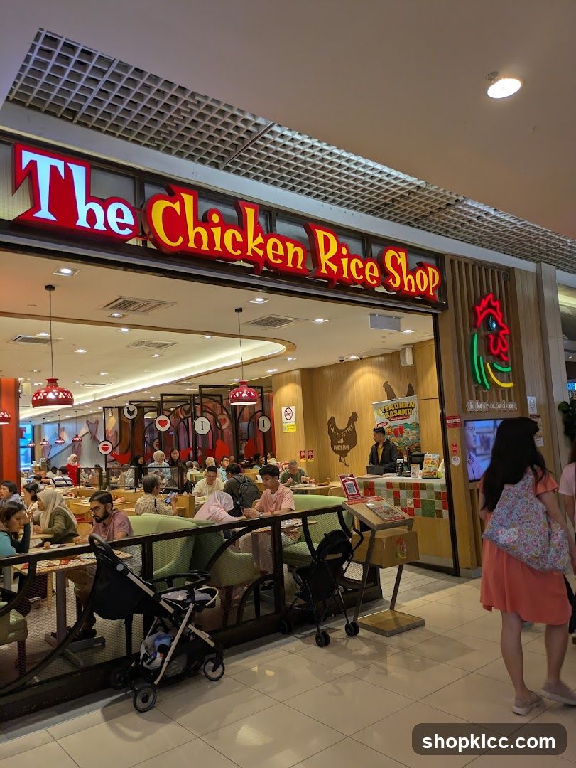 The exterior of a restaurant called "The Chicken Rice Shop" is visible. The restaurant has a prominent red and yellow sign with the name of the establishment. Inside, customers are seated at tables, and a staff member is visible behind a counter. The interior is decorated with chicken-themed imagery.