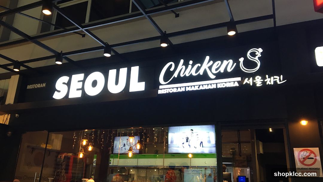 The exterior of a restaurant called Seoul Chicken. The sign is illuminated with white neon lights and features the words "SEOUL Chicken" in English and "서울치킨" in Korean. Below the main sign, it also says "RESTORAN MAKANAN KOREA". The restaurant has large glass windows displaying interior lights and screens showing what appear to be K-pop music videos. A "No Smoking" sign is visible on the right side of the entrance.