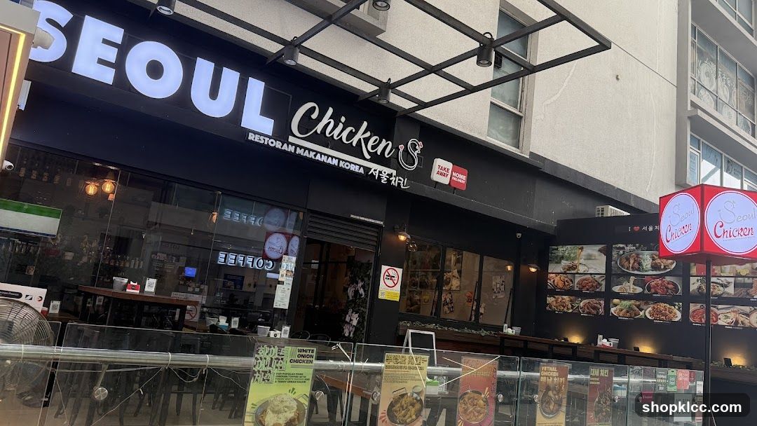 The exterior of a Seoul Chicken restaurant with large illuminated signage. The storefront features a black facade with the restaurant name "SEOUL Chicken" prominently displayed in white lettering. Below the main sign, smaller text reads "RESTORAN MAKANAN KOREA" and Korean characters. The entrance has large glass windows, and to the right, a red illuminated cube sign displays the "Seoul Chicken" logo. A display board with images of food is visible on the wall to the right of the entrance.