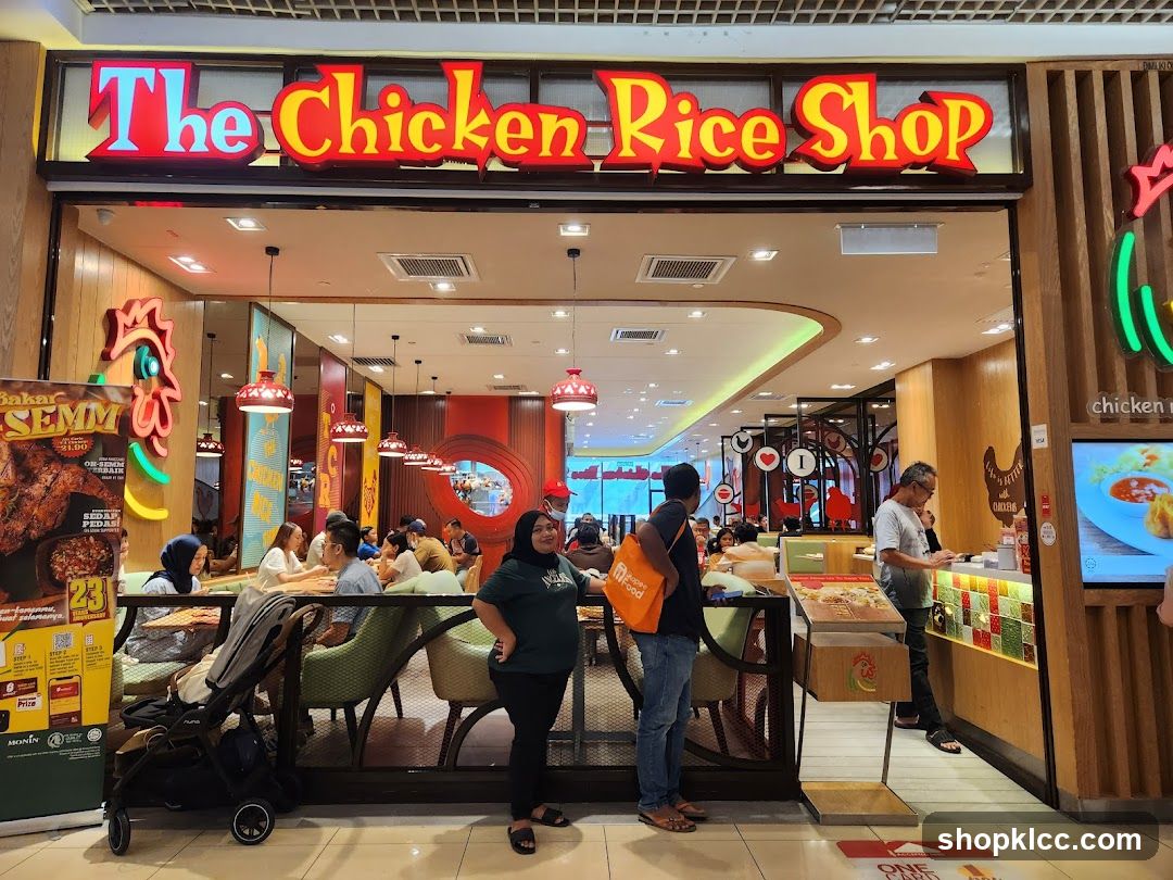 The exterior of a restaurant called The Chicken Rice Shop. The shop has a large, illuminated sign above the entrance. Inside, customers are seated at tables, and staff are visible behind a counter. There are decorative elements and food displays within the restaurant.