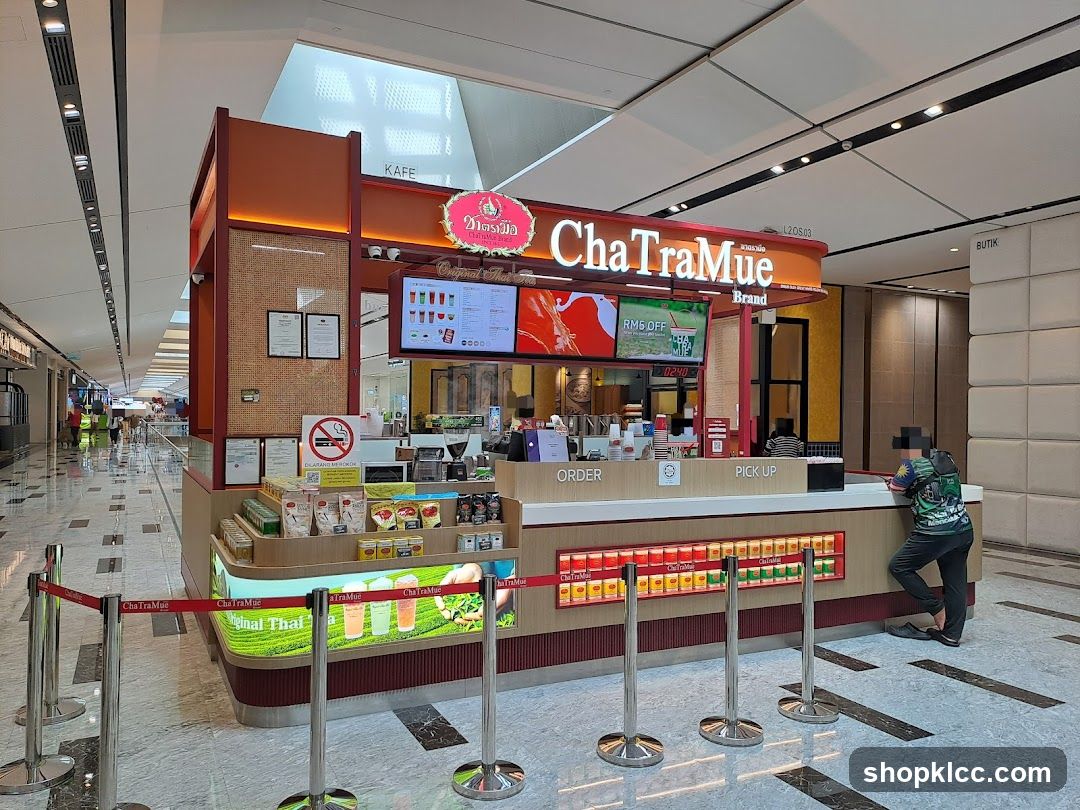 A ChaTraMue stall in a shopping mall, selling Thai tea and other beverages. The stall has a prominent sign with the brand name and logo. There are digital screens displaying promotions and menu items. The counter area has sections for ordering and pick-up. Various products, including tea packets and canned drinks, are displayed. A person is standing at the pick-up counter.
