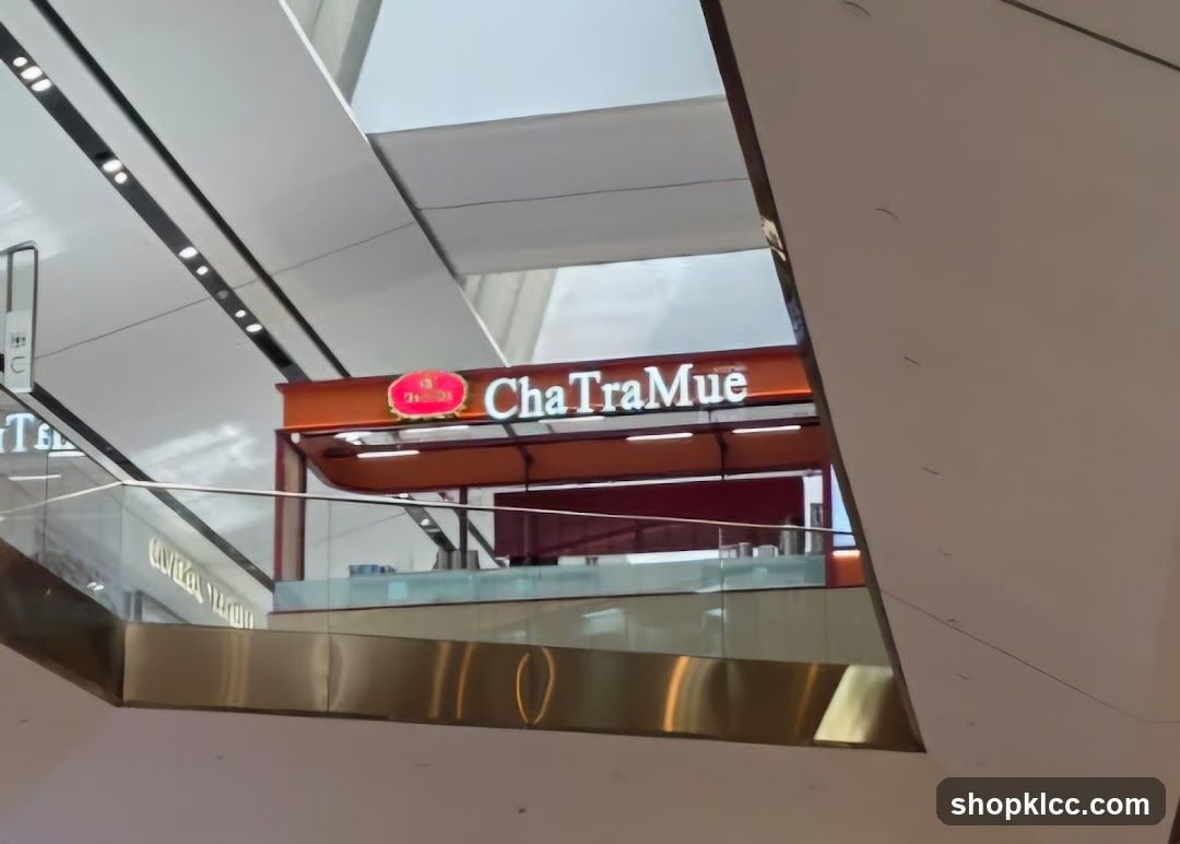A storefront for ChaTraMue, a tea shop, is visible in a modern indoor shopping mall. The shop features a prominent orange awning with the white illuminated text 'ChaTraMue' and a red oval logo. Below the awning, a counter area is visible with glass partitions. The surrounding architecture of the mall is sleek and contemporary, with white angled panels and metallic railings.