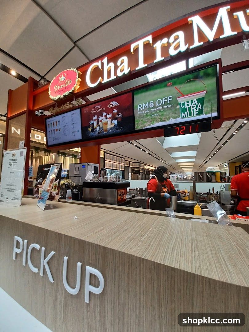 A view of the "ChaTraMue" shop front with a prominent "PICK UP" sign on the counter. Digital screens display menu items, promotions, and a timer. Staff members are visible behind the counter preparing drinks.