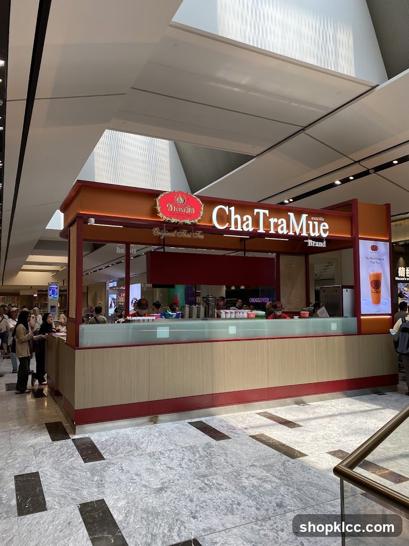 A brightly lit kiosk with the "ChaTraMue Original Thai Tea" logo prominently displayed. The kiosk has a modern design with wood-paneled lower walls and a red trim. Behind the counter, staff members are visible preparing drinks. A queue of customers is forming on the left side of the kiosk. The overall setting appears to be an indoor shopping mall or airport terminal.