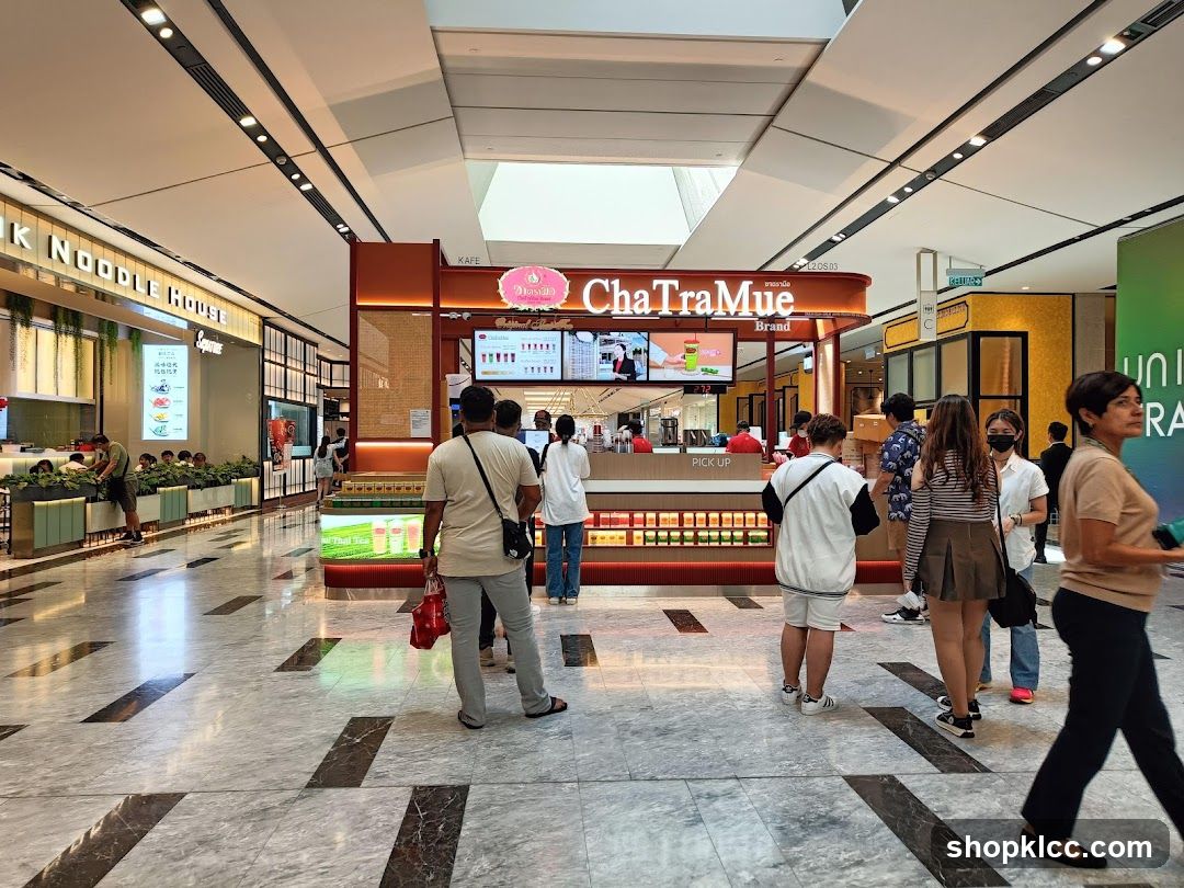 A ChaTraMue Brand shop front in a mall. The shop has a prominent orange and brown color scheme. There is a menu board displaying drinks and prices, and a pick-up counter. Several customers are visible in and around the shop.