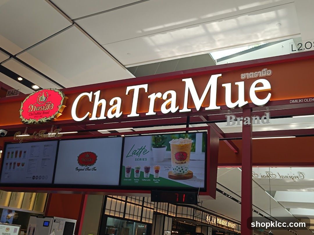 A storefront for ChaTraMue Brand, a beverage shop. The prominent signage displays the brand name "ChaTraMue" in large white letters against an orange background. Below the main sign, digital screens showcase menu items and product images, including "Original Thai Tea" and various "Latte Series" drinks like Cocoa Latte, Mocha Latte, Coffee Latte, and Black Tea Latte. The shop appears to be located in a mall or indoor shopping area, with other businesses visible in the background.