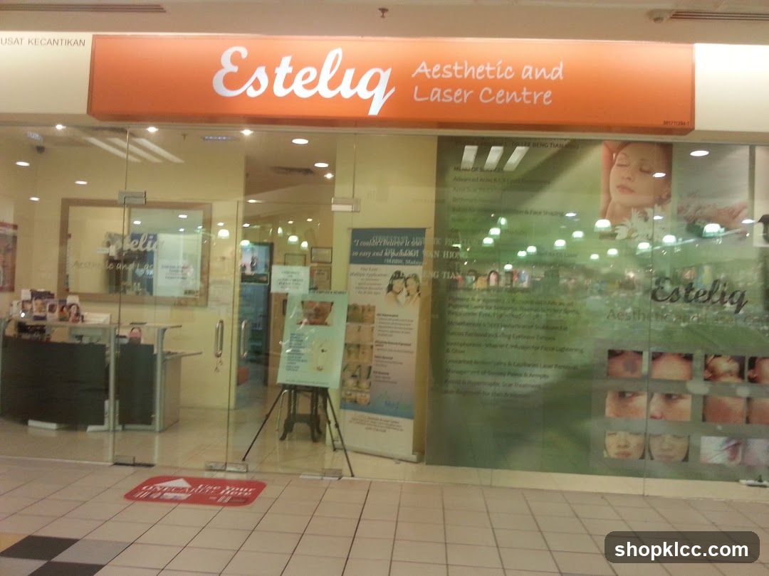 The exterior of an aesthetic and laser center with a large glass storefront. An orange sign above the entrance reads "Esteliq Aesthetic and Laser Centre". The glass windows display various advertisements and images related to beauty treatments, including before-and-after photos of skin treatments. Inside, a reception desk and seating area are visible.