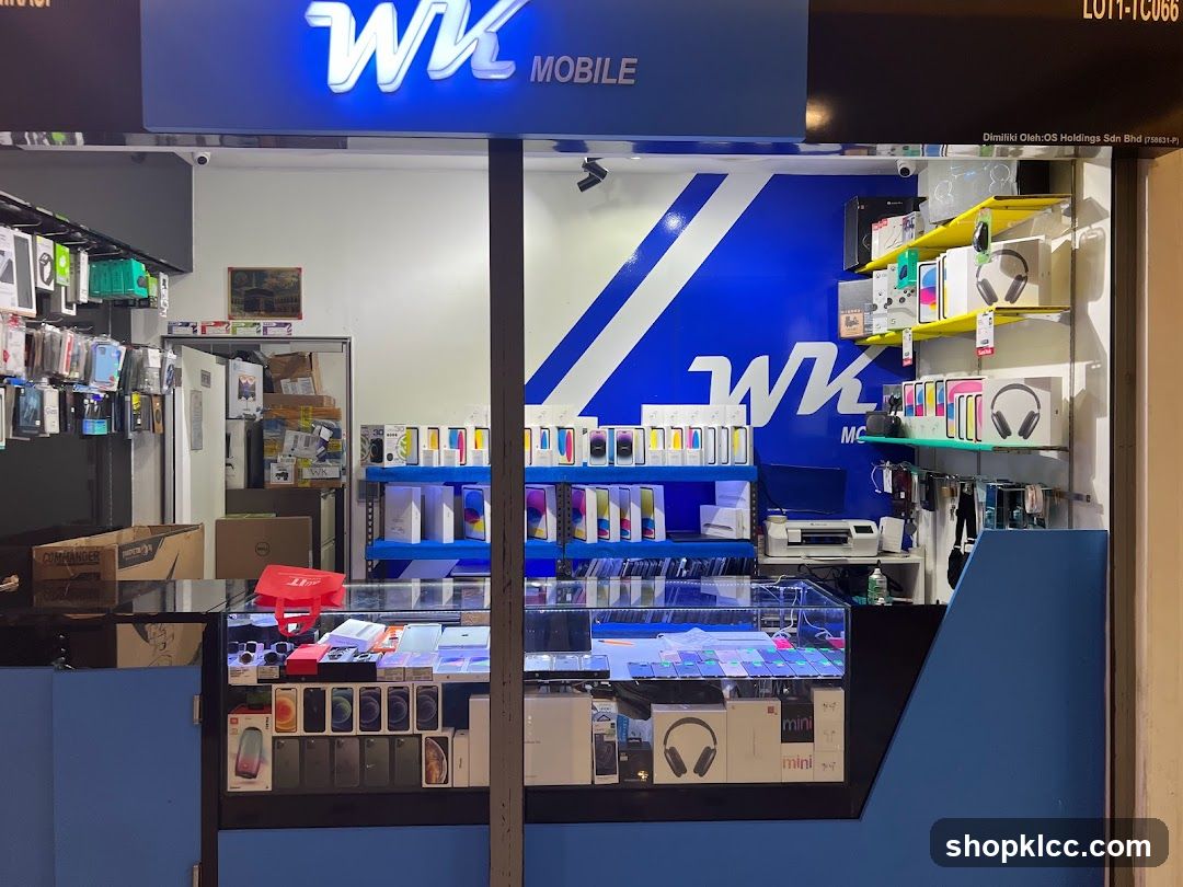 The exterior of a mobile phone store with a prominent blue and white sign displaying 'WK MOBILE'. The storefront features shelves stocked with various mobile phones, accessories, and headphones. A display counter showcases more phones and accessories. The interior is visible through the large glass windows.
