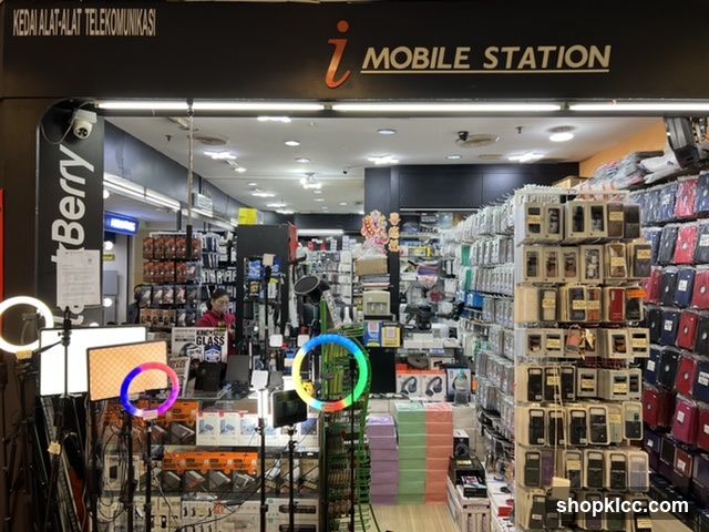 The exterior of a mobile phone accessories store. The storefront features a large sign with "i MOBILE STATION" and the text "KEDAI ALAT-ALAT TELEKOMUNIKASI" above it. The store is filled with various mobile phone accessories, including cases, screen protectors, and charging cables. Several ring lights and other photography equipment are displayed outside the entrance. A person can be seen inside the store.