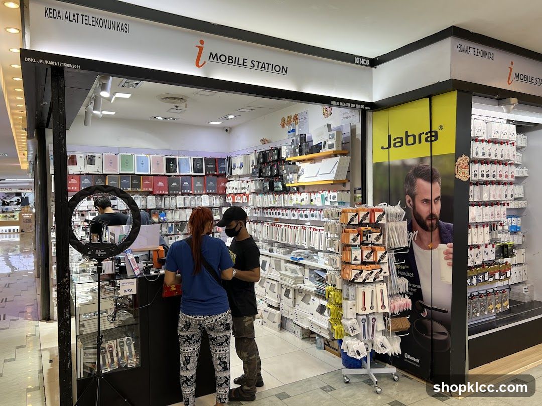 A telecommunications store with various mobile accessories displayed on shelves. A large Jabra advertisement featuring a man is visible on the right. Two customers are interacting with a staff member at the counter.
