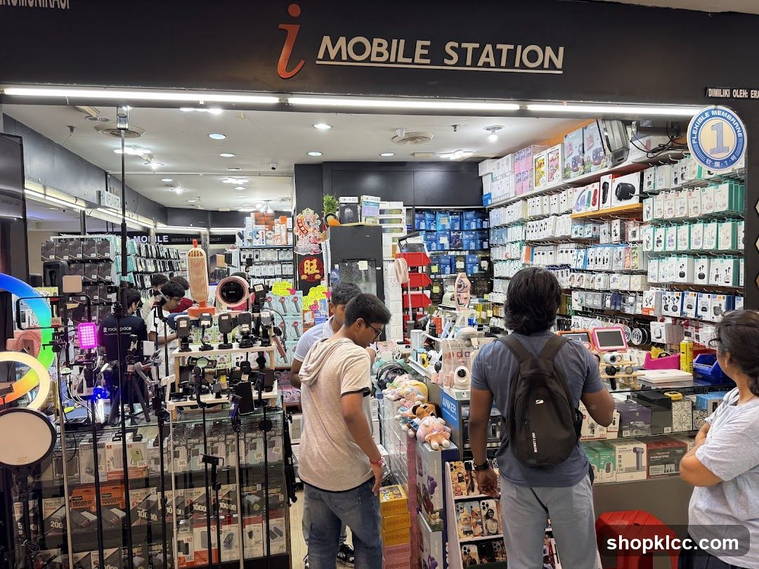 The exterior of a mobile accessories store called "i MOBILE STATION". The store is filled with various mobile accessories, including phone cases, chargers, headphones, and other electronic gadgets. Several customers are browsing the merchandise. The store's signage is clearly visible above the entrance.