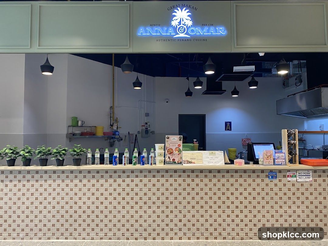 The storefront of a restaurant with a neon sign that reads "GERAI MAKAN AUNTIE PAK CIK ANNA OMAR AUTHENTIC PENANG CUISINE". The counter is decorated with small brown and beige tiles. On the counter, there are potted plants, bottles of water and soda, and various signs. A computer monitor and a wine rack are also visible.