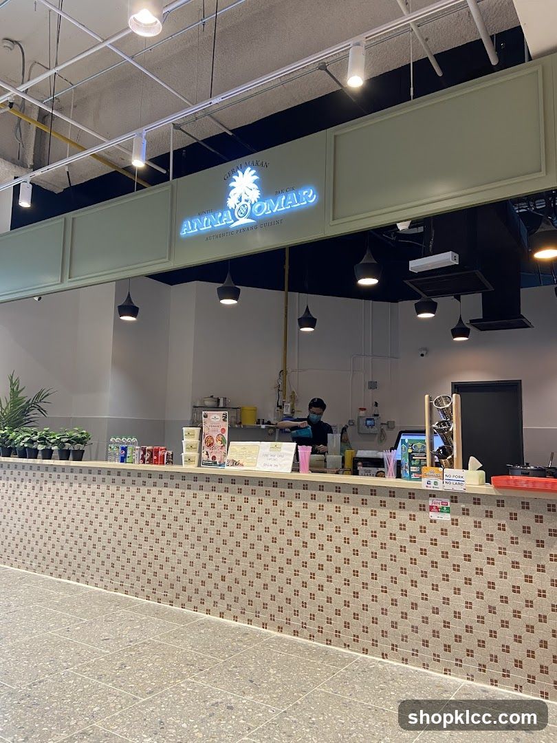 A view of the counter and signage of a food stall. The stall's name, "Auntie Anna & Omar Authentic Penang Cuisine," is displayed prominently in neon blue lettering above the counter. A staff member wearing a mask and apron is visible behind the counter, preparing food. The counter itself is decorated with small, square tiles.