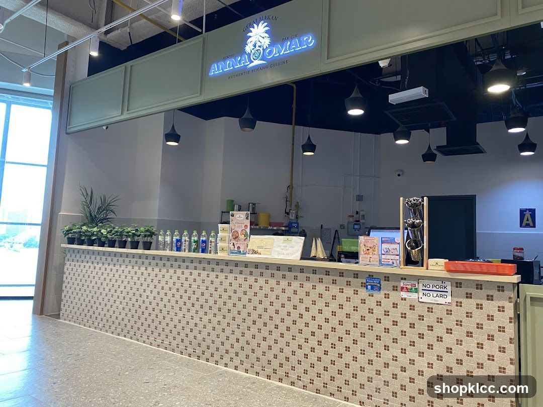 The exterior of a food stall with a sign that reads "GERAI MAKAN AUNTIE ANNA OMAR AUTHENTIC PENANG CUISINE". The counter is decorated with small brown and white tiles. There are plants and bottles of drinks on the counter. Signs indicating "NO PORK NO LARD" are visible.