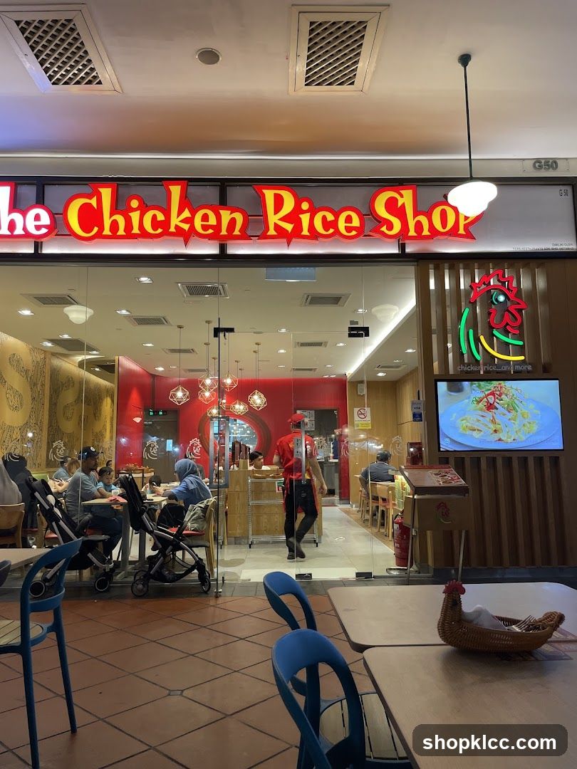 The exterior of a restaurant called "The Chicken Rice Shop". The sign is large and illuminated in red and yellow. Through the glass doors, customers can be seen dining inside. A digital menu board displaying a chicken rice dish is visible to the right of the entrance.
