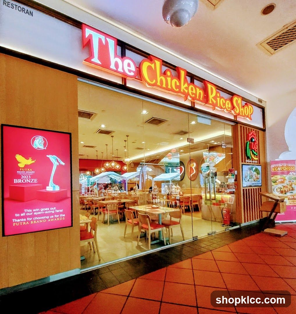 The exterior of a restaurant called "The Chicken Rice Shop". The restaurant has large glass windows and doors, revealing tables and chairs inside. A large red sign on the left displays a bronze award. The restaurant's name is prominently displayed in large, illuminated letters above the entrance.