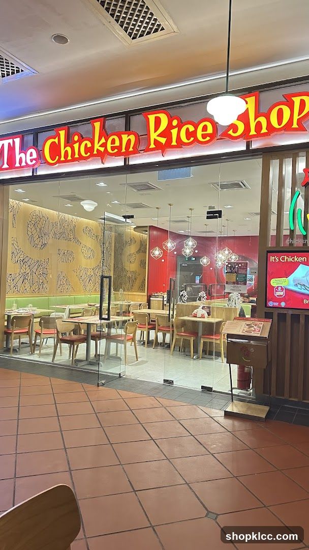 The exterior of a restaurant called "The Chicken Rice Shop". The entrance is through glass doors. Inside, there are tables and chairs, and the walls are decorated with a patterned design. A menu stand is visible outside the entrance.