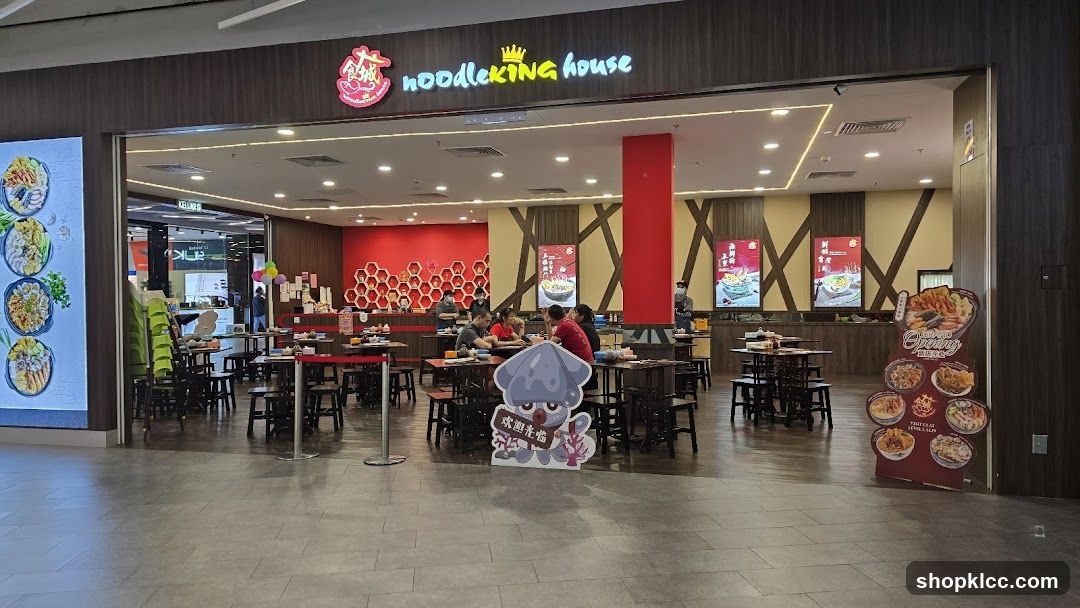 The exterior of a restaurant called Noodleking House. The restaurant has a wooden facade with the name and logo above the entrance. Inside, tables and chairs are set up for dining. There are also several posters displaying food items. A cartoon character cutout stands in front of the restaurant.
