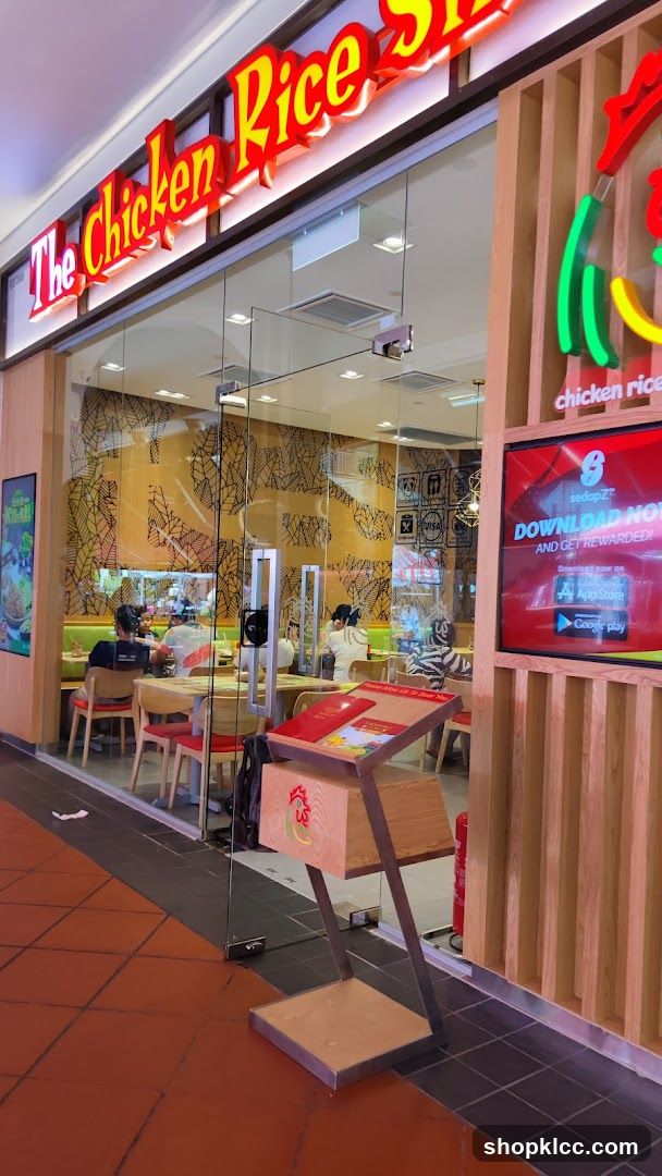 The exterior of a restaurant called "The Chicken Rice Shop". The entrance has large glass doors and windows. Inside, customers are seated at tables. A digital screen displays an advertisement for an app. A wooden stand with a menu is placed outside the entrance.