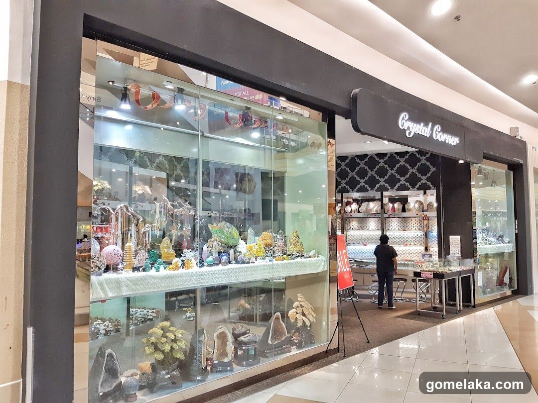 A storefront with a large glass display window showcasing crystals, minerals, and decorative items under bright lighting. The shop sign "Crystal Corner" is mounted above the entrance in white cursive lettering on a black background. Inside, shelves hold various crystal products and a customer is browsing near a counter. A red "SALE" sign stands outside the store.