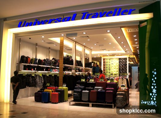 The exterior of a Universal Traveller store, showcasing a wide selection of luggage and travel accessories. The store features a prominent blue illuminated sign above the entrance. Inside, shelves are stocked with various backpacks and bags, while racks display jackets and clothing. A display of colorful suitcases is arranged in the foreground.