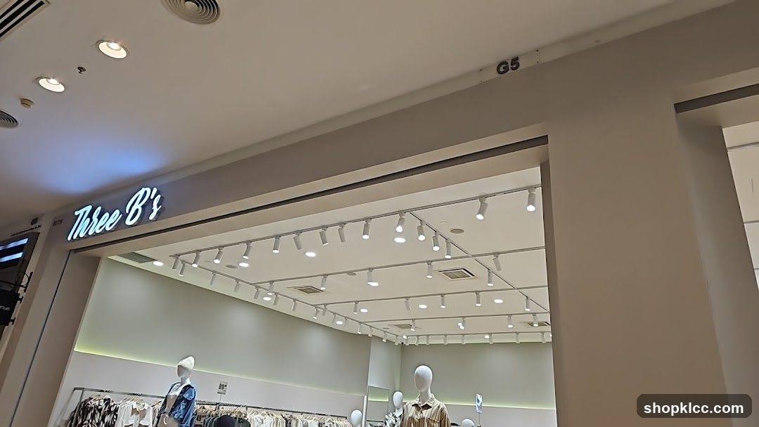 The image shows the exterior of a store with the name "Three B's" illuminated in a script font above the entrance. The store appears to be a clothing boutique, with mannequins visible inside displaying outfits. The interior is brightly lit with numerous track lights on the ceiling. A sign with "G5" is visible on the ceiling near the top right of the image.