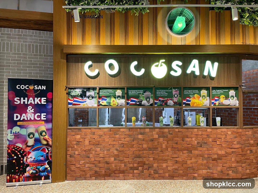 The storefront of a COCOSAN shop with a large illuminated sign that reads "COCOSAN" with a coconut graphic. Below the sign is a display window showcasing various drinks and promotional images. To the left of the storefront is a tall banner advertising "COCOSAN SHAKE & DANCE" with a cartoon cat graphic. The facade is decorated with wooden panels and brickwork.