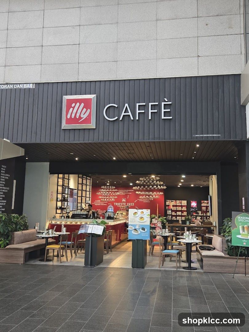 The exterior of an Illy Caffè coffee shop. The prominent signage above the entrance reads "illy CAFFÈ". The entrance opens to reveal a brightly lit interior with a red counter, seating areas, and displays. Tables and chairs are set up both inside and outside the shop. A seasonal special poster is visible to the right.