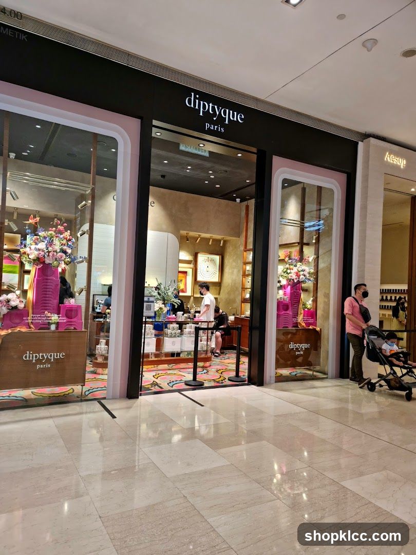 The exterior of a Diptyque Paris store, featuring large display windows showcasing floral arrangements and products. The store's name, 'diptyque paris', is prominently displayed above the entrance. The entrance is open, with a glimpse of the interior and staff. To the right, the adjacent store 'Aesop' is visible.