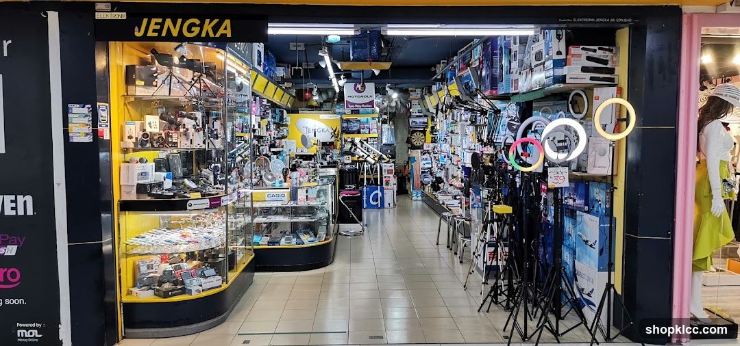 Store entrance with yellow and black signage displaying 'JENGKA' above the doorway. Interior is visible, showing shelves stocked with electronics, camera equipment, and lighting gear like ring lights on stands. A mannequin in a pink outfit is to the right. The floor is tiled, and ceiling lights illuminate the space.