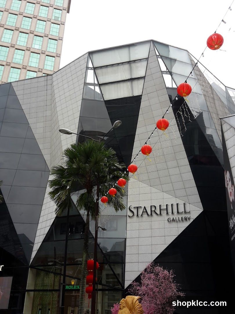 The exterior of a modern building with a geometric facade, featuring the signage "STARHILL GALLERY". Red lanterns are strung across the front, and a Rolex store is visible at street level. A palm tree stands in front of the building, and a decorative golden rooster and pink cherry blossoms are also present.