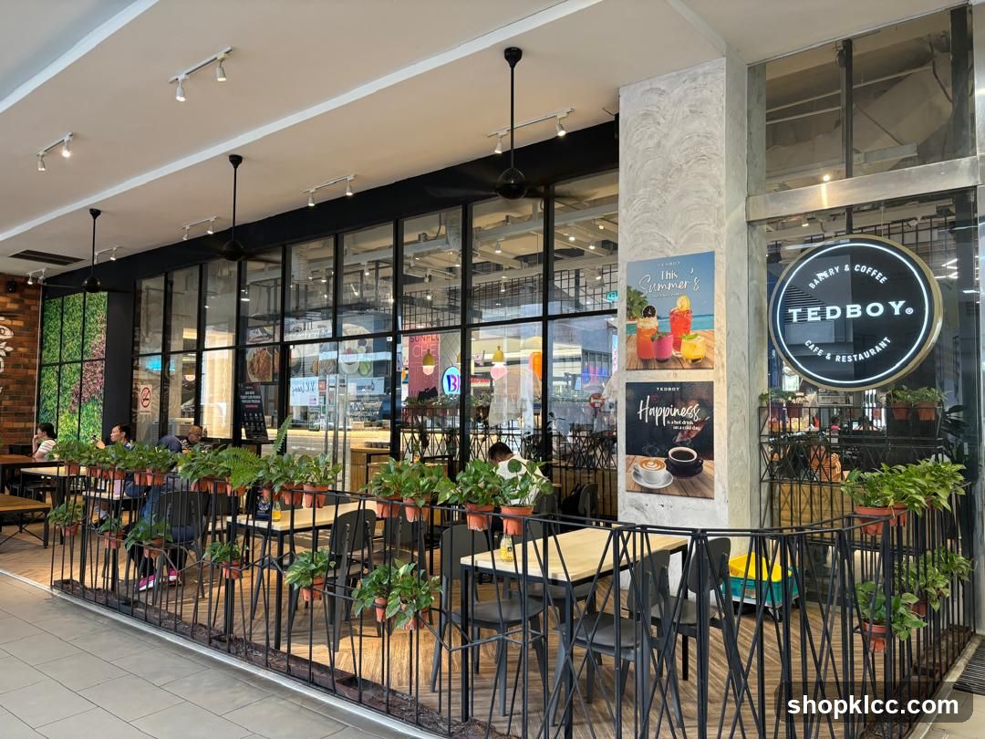 The exterior of a cafe and restaurant with a prominent circular sign that reads "TEDBOY. BAKERY & COFFEE CAFE & RESTAURANT". The cafe has large glass windows and doors, with an outdoor seating area separated by a black metal railing. Potted plants are placed along the railing and inside the cafe. Two posters are displayed on a pillar near the entrance. One poster advertises "This Summer's" drinks, and the other is titled "Happiness" and features a coffee cup.