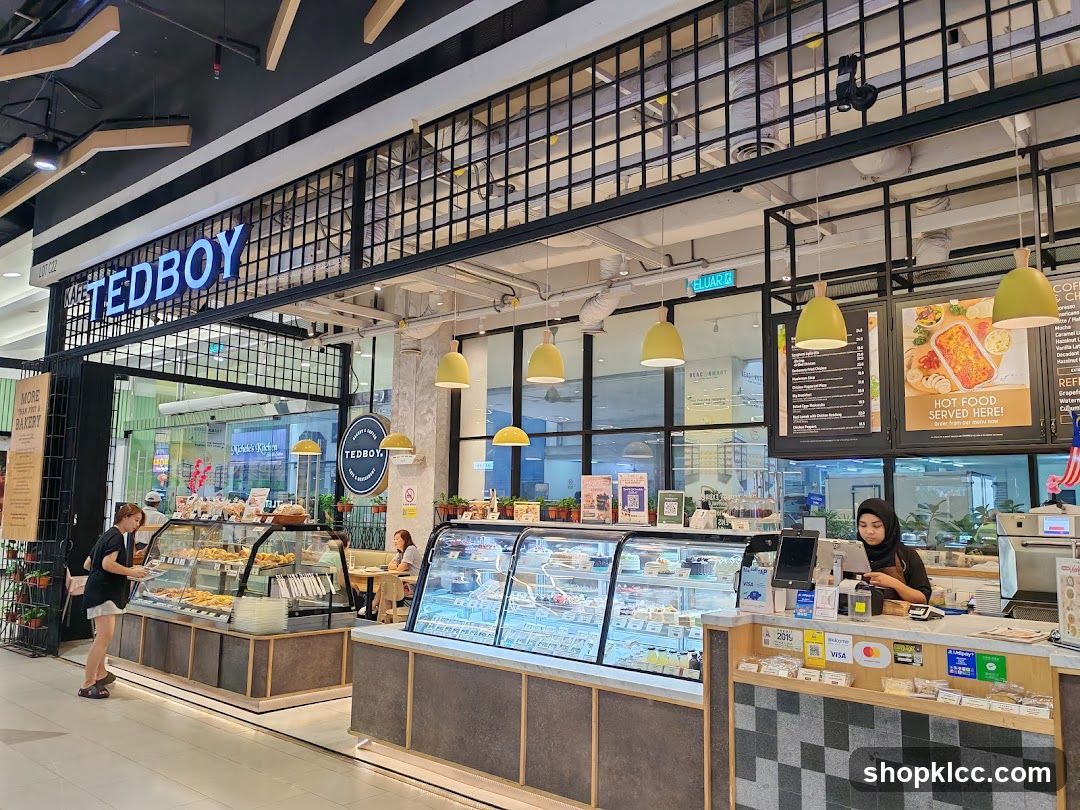 The exterior of a cafe and restaurant with a prominent "TEDBOY" sign. Display cases filled with pastries and cakes are visible, along with a counter where a staff member is serving customers. The cafe appears to be located inside a mall.