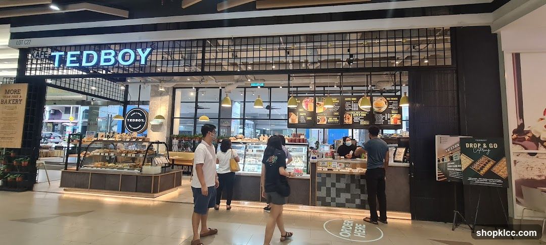 The exterior of a cafe called Kafe Tedboy, with a prominent sign above the entrance. Inside, display cases are filled with baked goods, and a counter serves customers. Menu boards are visible, displaying various drinks and food items. People are seen ordering and waiting.