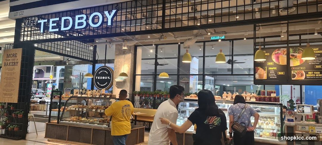 The exterior of a cafe with large glass windows displaying baked goods and a prominent "TEDBOY" sign above the entrance. A wooden sign to the left reads "MORE THAN JUST A BAKERY". People are seen inside and near the entrance.
