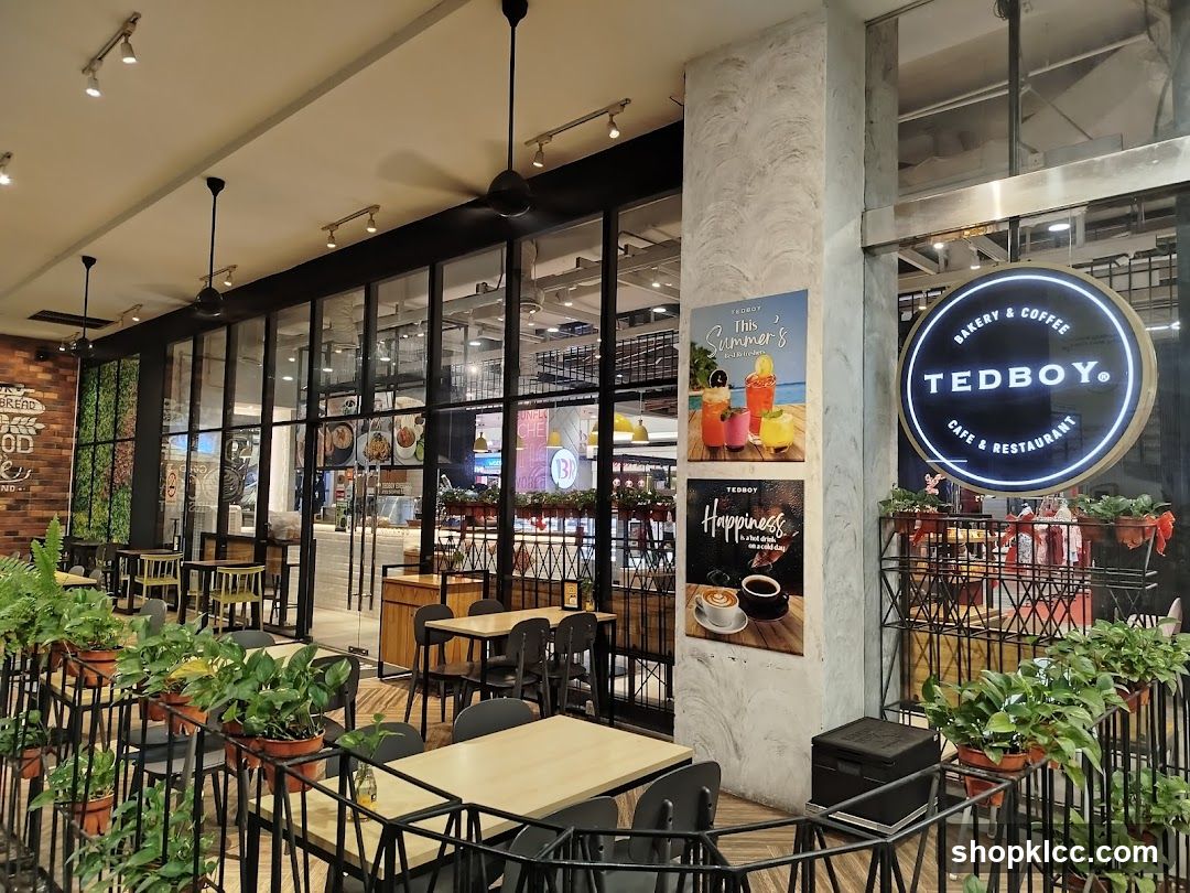 The exterior of a Tedboy Bakery & Coffee Cafe & Restaurant. A circular sign with the Tedboy logo is visible. There are tables and chairs set up outside the cafe, with plants decorating the area. Posters advertising drinks and coffee are displayed on a pillar.