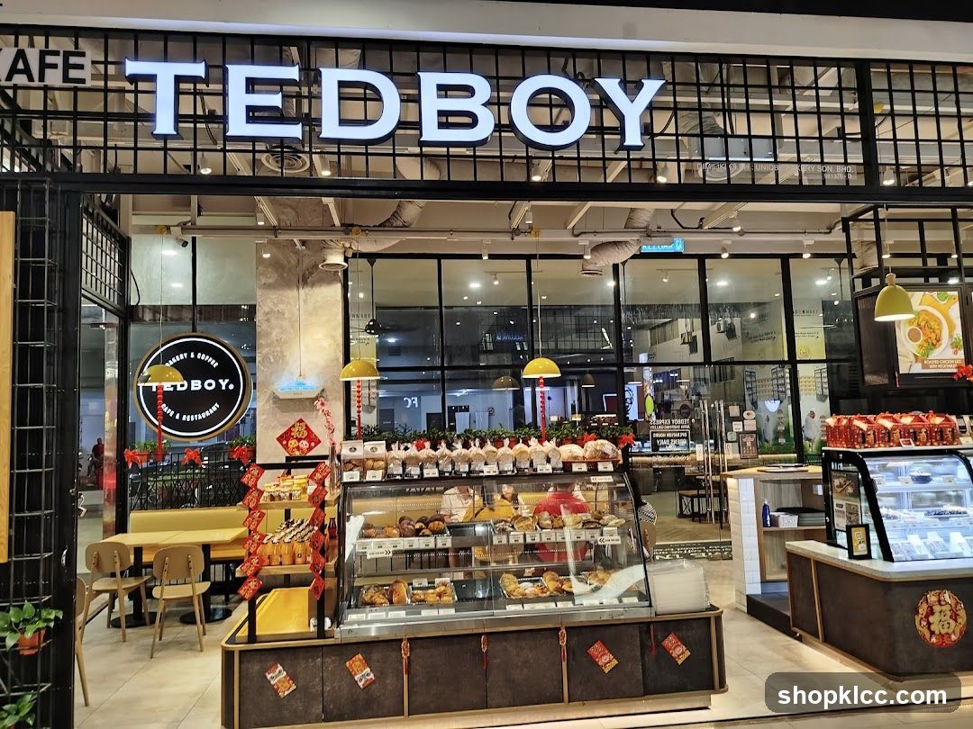 The exterior of a cafe with a large sign that reads "TEDBOY". The cafe has a display of baked goods and pastries in glass cases. There are also tables and chairs visible inside.