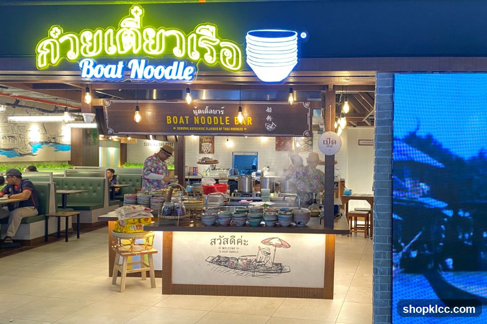 A brightly lit noodle bar with neon signage in Thai and English reading "Boat Noodle". The storefront features a counter with many bowls, cooking stations, and staff preparing food. Seating areas with green booths are visible in the background. An "Open" sign is displayed.