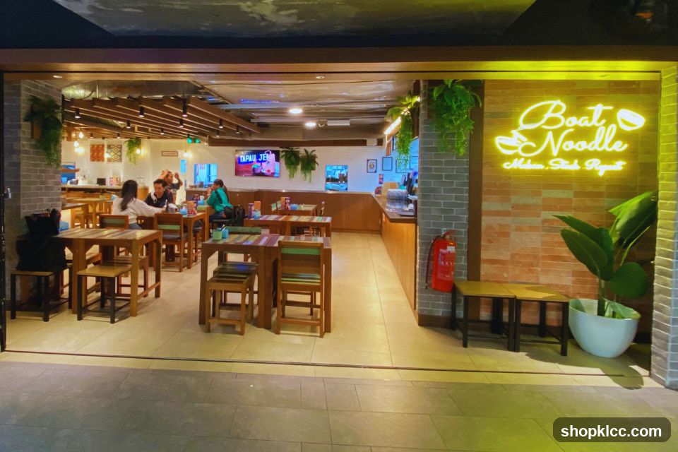 The exterior of a restaurant with a neon sign that says "Boat Noodle" and a tagline below. The restaurant has an open-air entrance with tables and chairs visible inside. There are also plants and a fire extinguisher near the entrance.