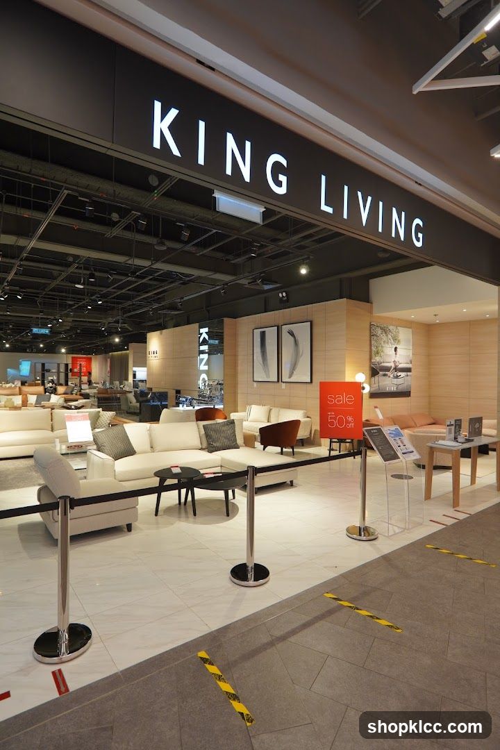 The image shows the entrance to a King Living furniture store. The store's name is prominently displayed in large white letters on a dark background above the entrance. Inside, various sofas and living room furniture are displayed. A red sign indicating "sale 50% off" is visible. The floor is tiled, and stanchions with black ropes guide visitors. Yellow and black hazard tape is present on the floor, suggesting a queueing area or safety markings.