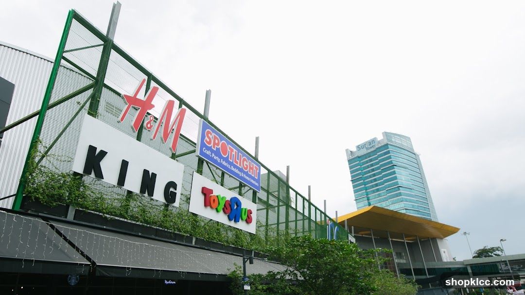 A low-angle exterior shot of a shopping complex featuring prominent signage for H&M, Spotlight, King, and Toys R Us. The signs are mounted on a green mesh fence above the shops. In the background, the Surian Tower, a modern glass skyscraper, is visible. The foreground shows the awnings and decorative lights of the shops below.