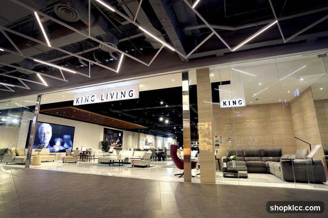 The exterior of a King Living furniture store is shown, with large glass windows displaying various sofa sets and living room arrangements. The store's name, "KING LIVING," is prominently displayed in large white letters on a dark background above the entrance. Inside, modern lighting fixtures illuminate the showroom, which features a wide selection of sofas, chairs, coffee tables, and other home furnishings. A large screen on the left displays a portrait of a man.