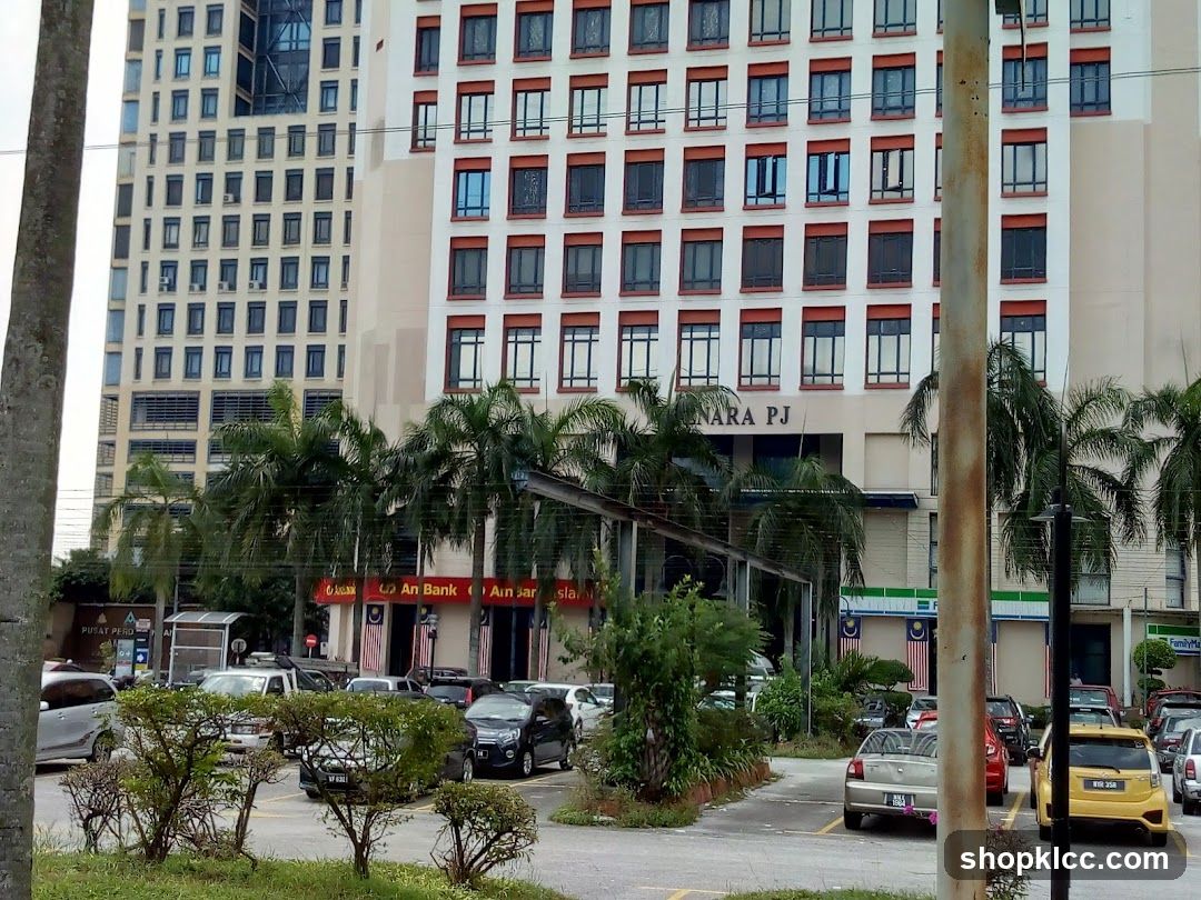 A wide shot of a building facade with multiple windows and balconies. The lower level features a prominent red sign that reads "An Bank" with Malaysian flags displayed. Palm trees are in front of the building. Several cars are parked in the foreground.