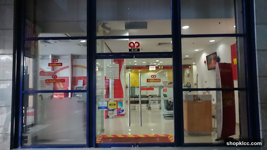 The exterior of an AmBank branch with large glass windows and doors. The AmBank logo and "AmBank Islamic" are visible on the windows and inside the branch. Inside, counters, chairs, and signage are visible.